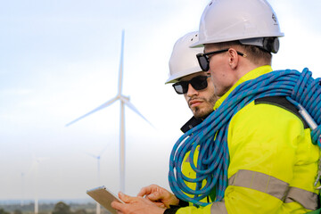 Workers in high visibility jackets examine a wind turbine farm, one pointing towards the turbines while the other holds a mobile device. The scene emphasizes renewable energy technological inspection.