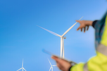A blurred figure holding a tablet observes large wind turbines in the background under a clear blue sky, symbolizing renewable energy, modern technology, and sustainable environmental solutions.