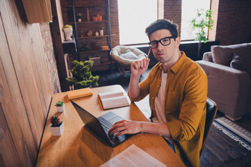 Young attractive man with glasses sitting at desk in modern loft living room, contemplating thoughtfully while using laptop