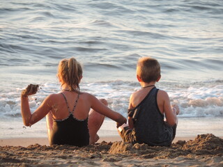 Niña y niño rubios, sentados frente al mar jugando con la arena