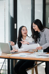 Two businesswomen discussing work at a desk with a laptop, showcasing teamwork and collaboration in a modern office setting.