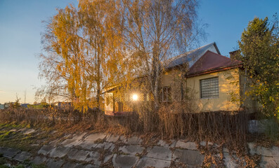 Dom na małym wzgórzu nad rzeką wśród brzóz ze złotymi jesiennymi liśćmi oświetlonymi zachodzącym słońcem. Mały dom wśród drzew na umocnionym brzegu małej rzeki (strumienia?!) wśród drzew . © Grzegorz