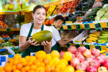 Positive young saleswoman holding fresh melon during offering fresh fruits in greengrocery store