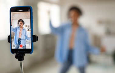 Modern Content Creator. Positive African American young lady in headset filming video blog for social network, using cellphone on tripod for broadcast in living room, selective focus on device screen