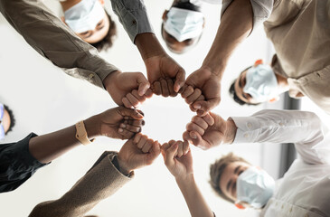 Success And Teamwork Concept. Below view of young diverse group of business people in protective medical masks making fist bump standing in circle. Workers do fist pump together celebrating good deal