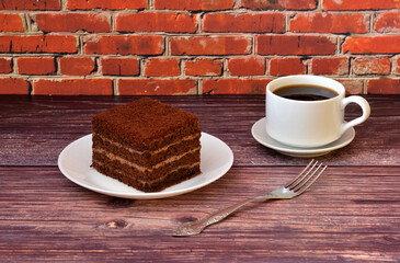 A piece of sponge chocolate layer cake on a white plate and a cup of hot black coffee on a wooden table, a fork nearby.