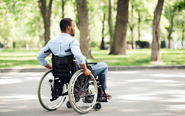 Fototapeta premium Thoughtful young black guy in wheelchair having walk at urban park, copy space. Millennial African American man with physical disability spending time outdoors, enjoying nature