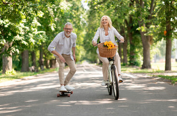 Modern Pensioners. Happy Senior Couple Riding Bike And Skateboard In Park, Cheerful Mature Man And Woman Having Fun Outdoors Together, Enjoying Active Lifestyle On Retirement, Copy Space