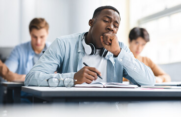 Tired black student yawning sitting at desk in classroom. African American youth exhausted from getting ready for test or writing coursework, feeling sleepy during lecture in highschool. Lack of sleep
