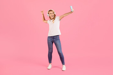 A young woman enjoys her time, dancing and taking a selfie while wearing headphones. The vibrant pink background enhances her cheerful mood and lively expression in this playful moment.