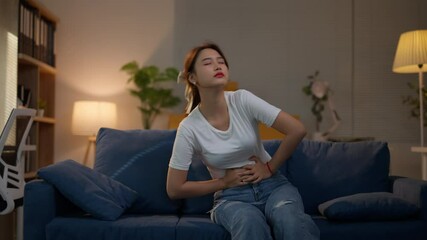 Young woman is experiencing intense stomach pain while sitting and then lying on her couch at night - Powered by Adobe