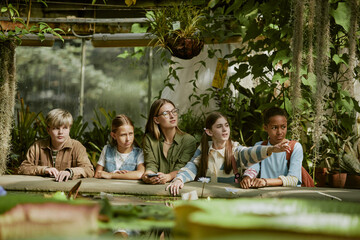 Young female teacher and group of teenagers discussing plants during tour in botanical garden greenhouse, copy space