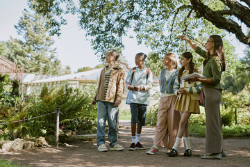 Young female botanical garden worker showing rare plants to group of teen students during tour,...