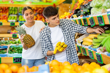 Married couple picks and buys ripe bananas and pineapple in vegetable section of grocery supermarket