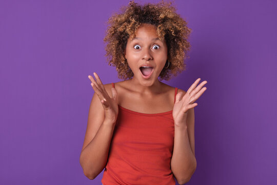 Young African American female with curly hair shows excitement and surprise with wide eyes and raised hands against a vibrant purple background. Her joyful expression captures a moment of pure delight