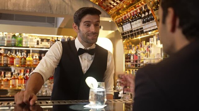 Smart bartender serving drink gin fizz with lemon sliced to VIP guest while waiting friend at luxurious counter bar. Barman providing excellent service at vibrant nightlife party atmosphere. Vinosity.