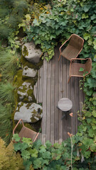 Fototapeta premium Overhead view of a wooden deck with two wicker chairs, a small round table, and several mossy rocks surrounded by lush green vegetation.