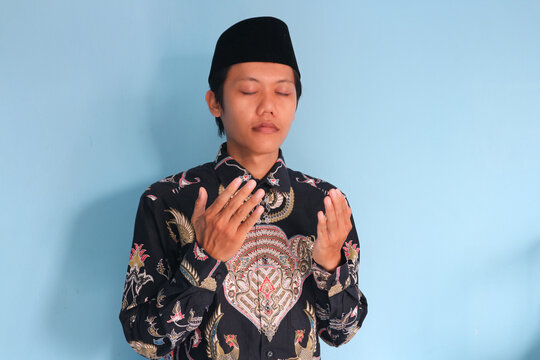 Young asian muslim man wearing a batik shirt and songkok raising hands while praying to Allah SWT