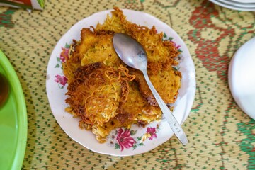 A top view of a plate of fried noodles with a fried egg on top, served on a traditional Indonesian woven mat. A spoon is placed on top of the fried egg.