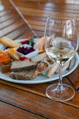 A glass of white wine on a wooden slatted table and an appetizer on a white plate, bread, cheese, fruit, sauce. Wine tasting, brunch