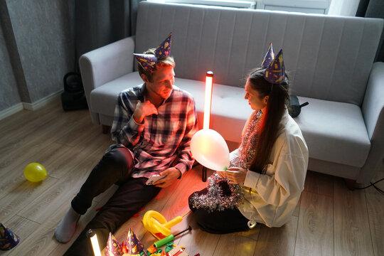 Couple enjoys a festive celebration on the floor, wearing party hats and surrounded by colorful decorations and balloons in a cozy indoor ambiance
