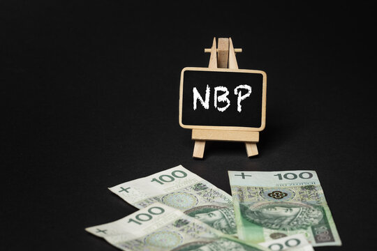 Black wooden writing board on a black background with the inscription "NBP", in the background scattered Polish złoty PLN banknotes (selective focus)