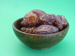 A wooden bowl filled with plump, glossy dates symbolizes abundance, nourishment, and the sweetness of life. Against a vibrant green background, these dates represent both natural simplicity.