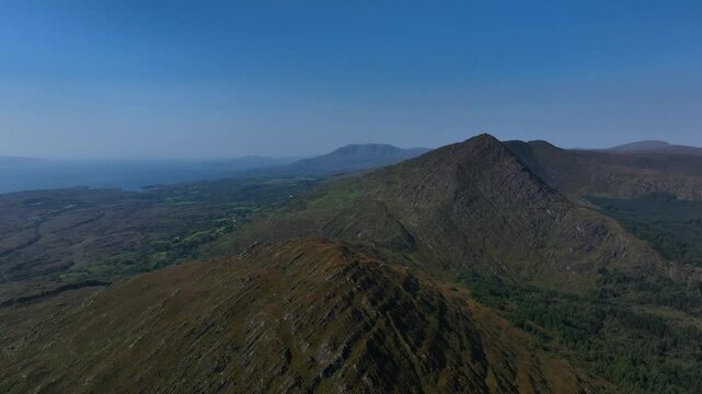 Sugarloaf, Caha Mountains, County Cork, Ireland, September 2024. Drone pulls backwards across the stunning mountainous peaks surrounded by a rugged craggy landscape under a clear blue sky.
