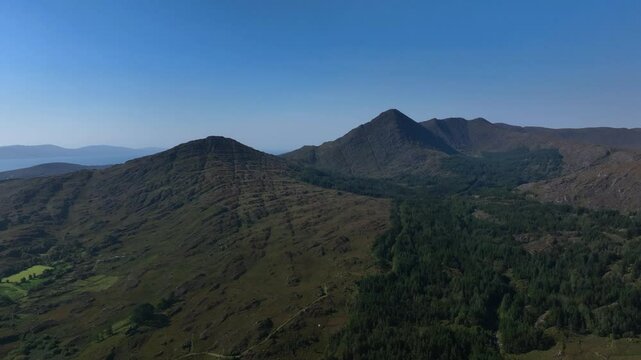 Sugarloaf, Caha Mountains, County Cork, Ireland, September 2024. Drone pulls backwards with a wide overview of the towering peaks surrounded by craggy fields and planted forest under a clear blue sky.