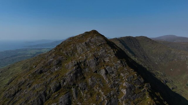 Sugarloaf, Caha Mountains, County Cork, Ireland, September 2024. Drone orbits counter clockwise around the majestic craggy summit under a clear blue sky with the North Atlantic Ocean in the distance.
