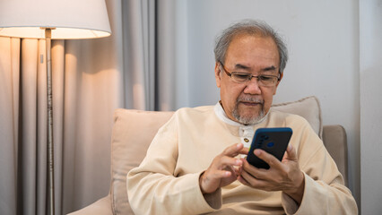 Asian old man engrossed reading a message in his mobile phone while sitting on a couch at house, Happy senior man sit in armchair relaxing with smartphone in living room at home, Technologies online