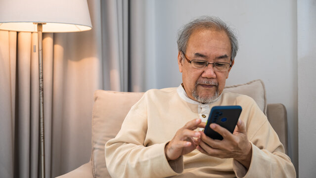 Asian old man engrossed reading a message in his mobile phone while sitting on a couch at house, Happy senior man sit in armchair relaxing with smartphone in living room at home, Technologies online