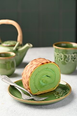 Pandan swiss roll or pandan roll cake placed on a green ceramic plate, accompanied by green ceramic cup and teapot ornaments - blurred background.