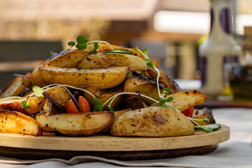 Crispy baked potato wedges garnished with microgreens and cherry tomatoes on wooden plate, side view. Outdoor dining, backyard dinner.