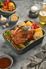 A whole grilled roast chicken served with slices of corn, tomatoes, carrots, mustard greens, and green beans - placed on a textured gray table, perfect for thanksgiving, breakfast, lunch, or dinner.