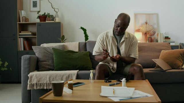 Middle-aged Black man with diabetes sitting on sofa in living room, measuring his blood sugar level, looking at medical device screen and shaking his head in disappointment