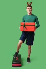 Young sporty man in reindeer horns holding Christmas gift box on blue background