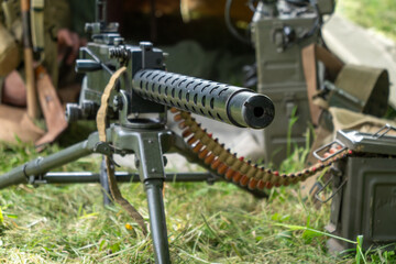 Black machine gun with a long barrel is on a grassy field. The gun is surrounded by a lot of ammunition