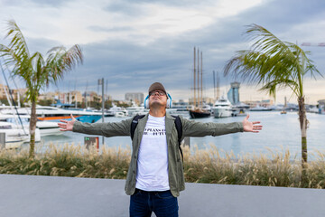 Happy man enjoying music with headphones, arms outstretched, at the city's marina