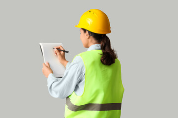 Young female construction worker with notebook and pen on grey background. Woman in male profession concept