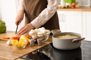 Cooking pot with soup on stove against woman cutting vegetables in kitchen, closeup