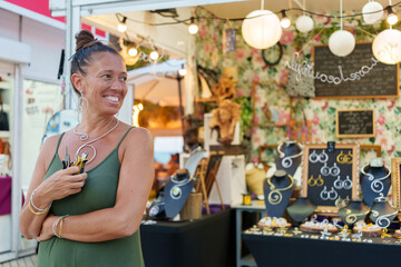 Woman artisan holding pliers smiling in her jewelry stall at a crafts market