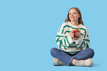 Beautiful young woman with glass cup of hot mulled wine sitting on blue background