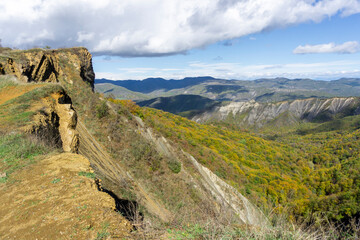 Gorge covered with autumn bushes and trees illuminated by the sun. Bare rocks are visible. Mountains, hills and sky with clouds in the background
