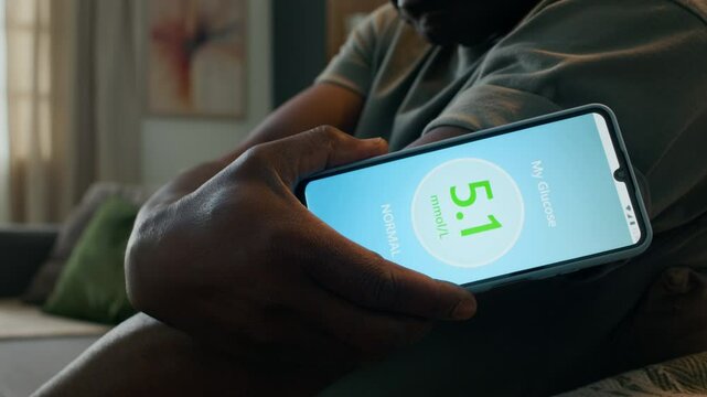 Close-up view of Black man holding smartphone displaying normal glucose reading from continuous glucose monitor sensor, then checking result in modern app while managing diabetes at home