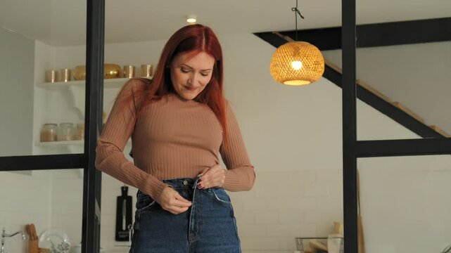 Irritated charming redhead mature woman with weight problems trying to button up and zip up tight jeans, expressing despair and disappointment while standing in domestic room.
