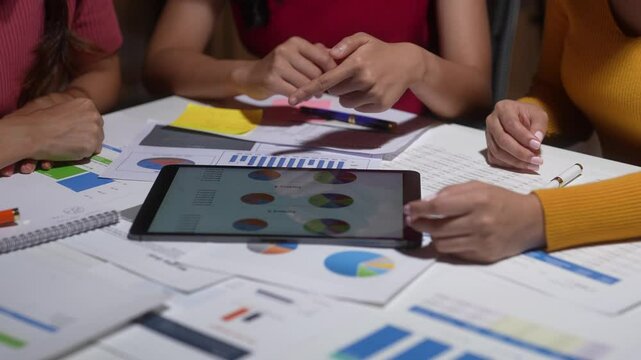 A woman is pointing at a tablet with a pie chart on it. The chart is on a table with other papers and a notebook. The woman is looking at the tablet with a serious expression