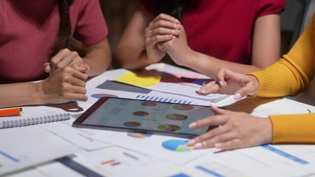 Three women are sitting at a table with a tablet in front of them. They are looking at the tablet and seem to be discussing something. The tablet is displaying a graph or chart