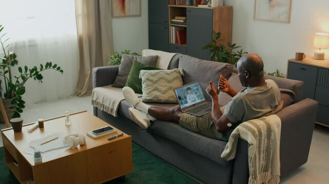High angle view of African American man resting on sofa in cozy living room, showing glucose meter sensor on arm and consulting about diabetes treatment with doctor via video call on laptop