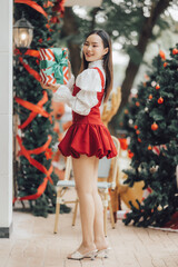 Christmas Cheer: A young woman smiles radiantly as she holds a beautifully wrapped gift, surrounded by festive Christmas trees. The scene captures the joy and excitement of the holiday season. 